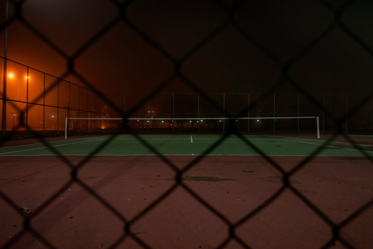 A tennis court captured through a wire fence at night with atmospheric lights.